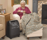 Old lady sitting on her sofa holding a cup of tea while using the Caire Visionaire 5 Stationary Oxygen Concentrator. Shop now at Zone Medical.